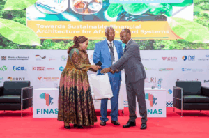 Dr. Charity Mutegi (l) and Tharaka University Chancellor, Prof. Ratemo Michieka (c) presenting a gift to Livestock Development PS, Jonathan Mueke (r) during the opening ceremony of FINAS 2026 at KICC, Nairobi