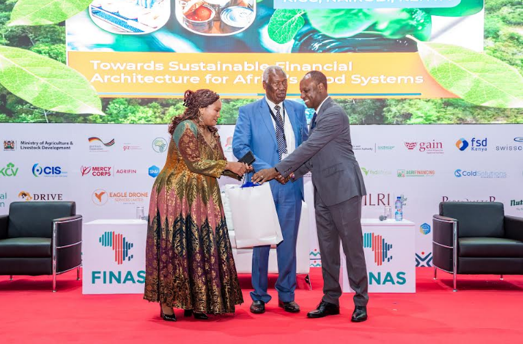 Dr. Charity Mutegi (l) and Tharaka University Chancellor, Prof. Ratemo Michieka (c) presenting a gift to Livestock Development PS, Jonathan Mueke (r) during the opening ceremony of FINAS 2026 at KICC, Nairobi